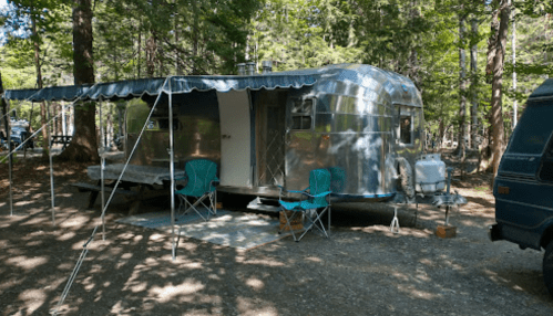 A shiny silver Airstream trailer parked in a wooded area, with two green chairs and a small table outside.