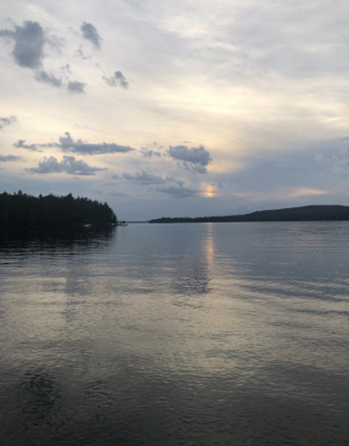 A calm lake at dusk, reflecting clouds and trees under a cloudy sky.