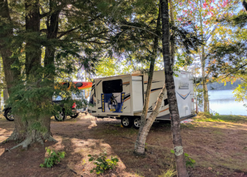 A camper parked among trees by a serene lake, with a kayak on top and colorful foliage in the background.