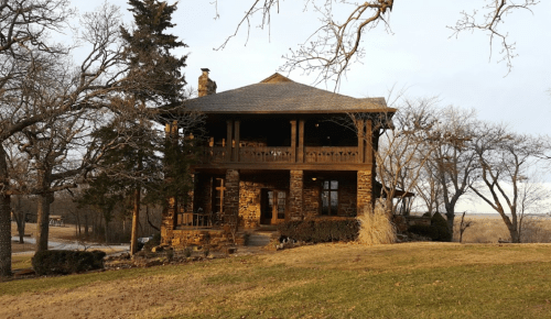 A rustic two-story house with a porch, surrounded by trees and a grassy lawn under a cloudy sky.