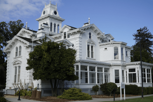 A large, white Victorian-style house with a tower, surrounded by greenery and a clear blue sky.