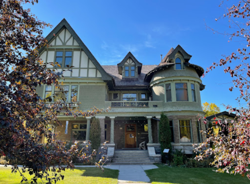 A large, historic house with a mix of architectural styles, surrounded by trees and a clear blue sky.