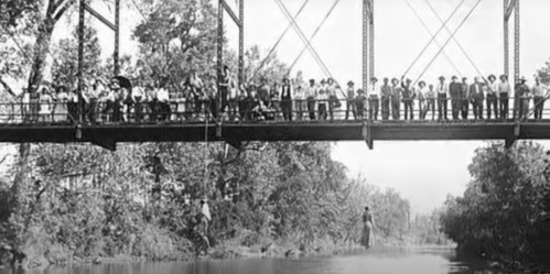 A historic black-and-white photo of a large group of people standing on a bridge over a river, surrounded by trees.