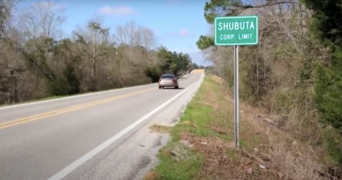 A road sign indicating the corporate limit of Shubuta, with a car driving on a rural road surrounded by trees.