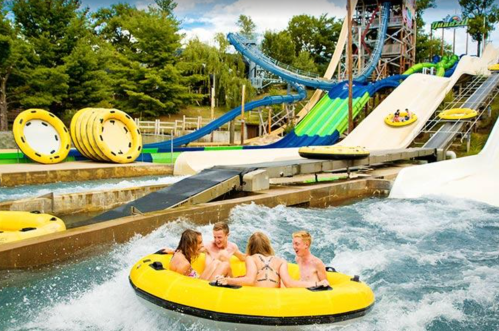 A group of four kids in a yellow inflatable raft enjoying a water slide at a water park on a sunny day.