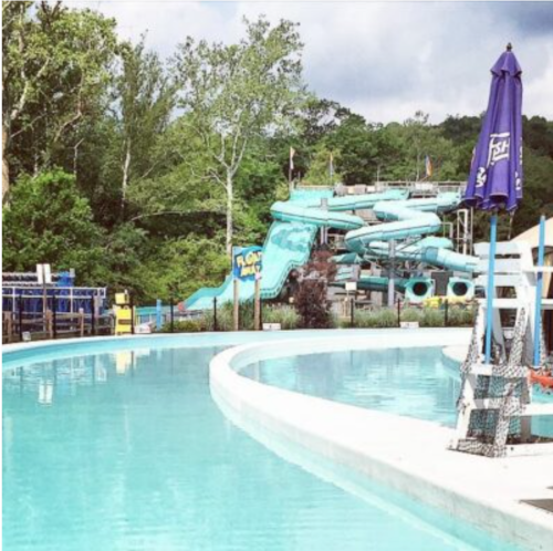 A clear pool with a winding water slide in the background, surrounded by trees and a cloudy sky.