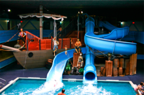 Indoor water park featuring a pirate ship, blue water slides, and people enjoying the pool.
