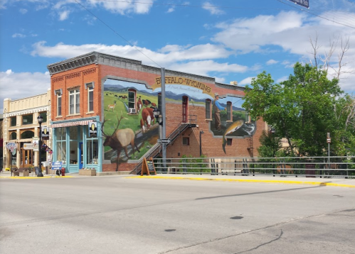 A brick building in Buffalo, Wyoming, featuring a colorful mural and a staircase, with blue skies and trees nearby.
