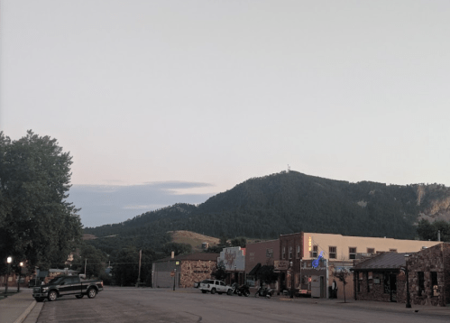 A quiet street scene with buildings and parked cars, set against a backdrop of rolling hills and a clear sky.