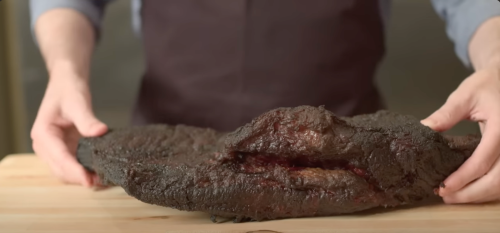 A person in an apron holds a large, smoked brisket over a wooden cutting board.