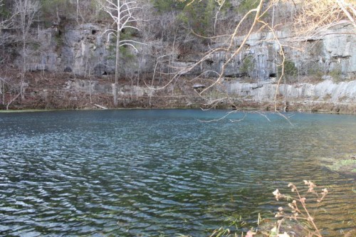 A serene body of water surrounded by rocky cliffs and sparse trees, reflecting a calm, clear sky.