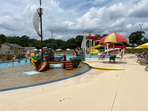 A water park scene featuring a pirate ship play structure, water slides, and people enjoying the pool under a cloudy sky.