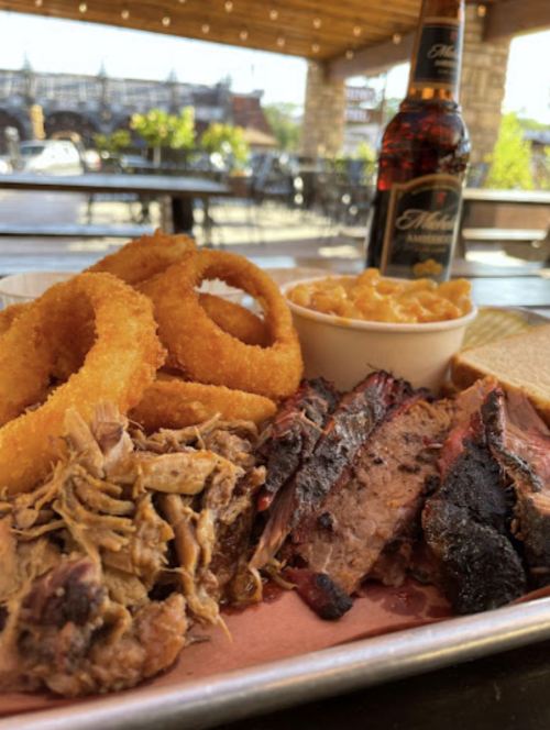 A platter of barbecue with pulled pork, brisket, onion rings, mac and cheese, and a beer in the background.