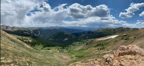 A panoramic view of a mountainous landscape with green valleys, rocky terrain, and scattered clouds in a blue sky.