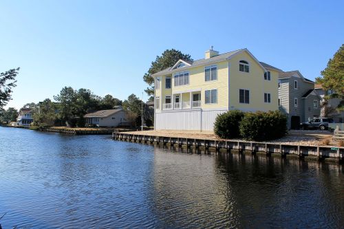 A yellow house by a calm waterway, surrounded by trees and other homes under a clear blue sky.