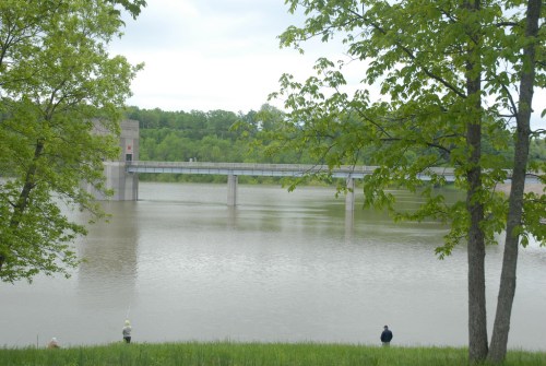 A serene river scene with a dam in the background, surrounded by green trees and a cloudy sky.
