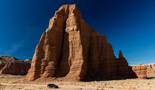 A towering rock formation rises against a clear blue sky, with a car parked nearby in a desert landscape.