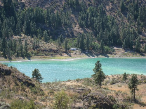 A serene lake surrounded by green trees and hills, with a small beach area and a few campers in the distance.