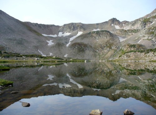 A serene mountain landscape reflected in a calm lake, surrounded by rocky terrain and patches of snow.