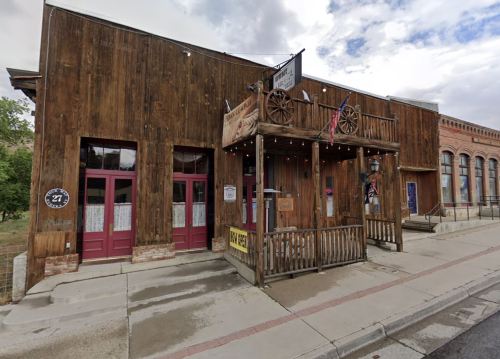 A rustic wooden building with a balcony, featuring red doors and western-themed decor, set against a cloudy sky.