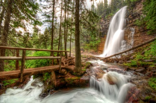 A serene waterfall cascades down rocky cliffs, surrounded by lush greenery and a wooden bridge over a flowing stream.