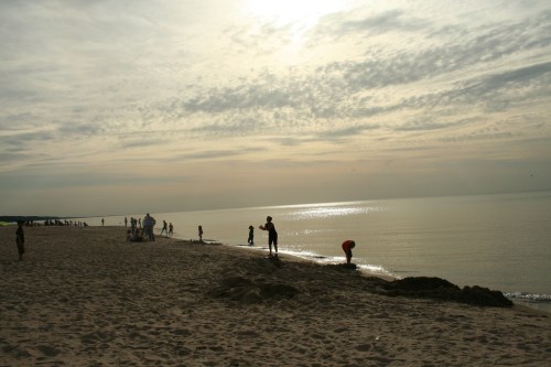 Silhouettes of people on a beach at sunset, with calm water and a cloudy sky in the background.