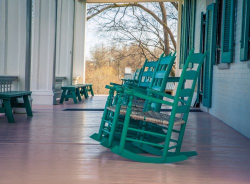 A row of teal rocking chairs on a porch with wooden flooring and trees in the background.