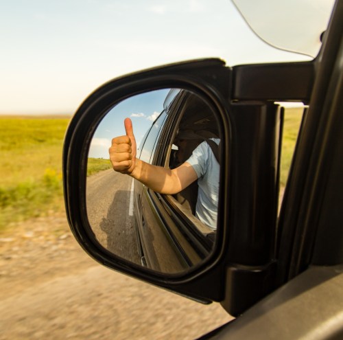 A hand giving a thumbs-up is reflected in a car's side mirror, with a scenic landscape in the background.