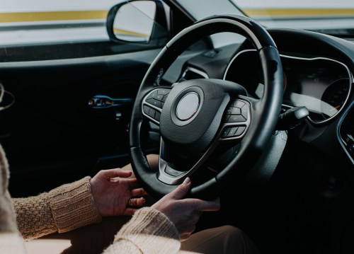 A close-up of hands gripping a car steering wheel, with a dashboard visible in the background.