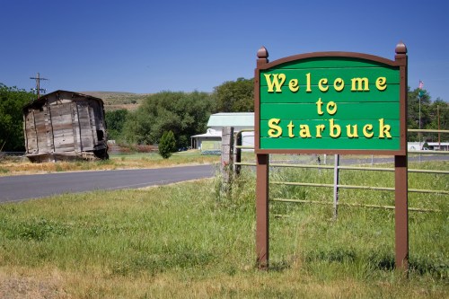 A green sign reading "Welcome to Starbuck" beside a road, with an old, leaning building in the background.