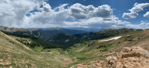 A panoramic view of a mountainous landscape with green valleys, rocky terrain, and scattered clouds in a blue sky.