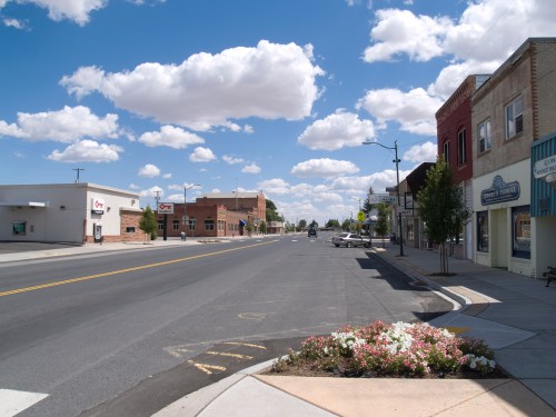 A quiet street scene with shops, trees, and fluffy clouds under a blue sky.