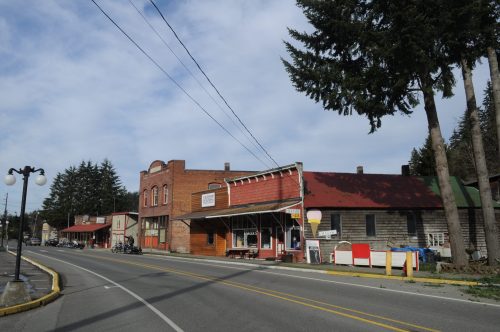 A small town street featuring brick buildings, shops, and trees under a cloudy sky.