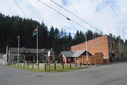 Historic buildings with a mix of brick and wood, surrounded by trees and a flagpole, in a small town setting.