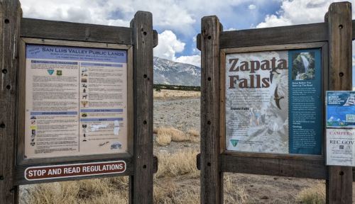 Information signs about Zapata Falls and regulations for San Luis Valley Public Lands, with mountains in the background.