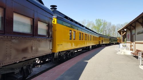 A row of vintage trains, including a bright yellow passenger car, at a train station surrounded by trees.