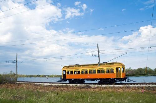A vintage yellow and orange trolley car on tracks beside a lake, with blue skies and clouds in the background.