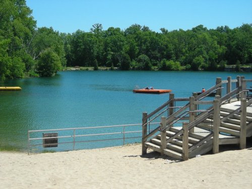 A sandy beach leads to a calm lake surrounded by green trees, with a kayak on the water and steps leading down to the shore.