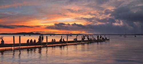 Silhouetted figures on a dock at sunset, with boats in the water and vibrant clouds reflecting in the calm sea.