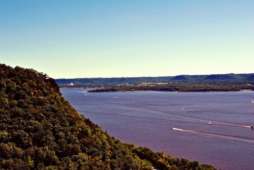 A scenic view of a river winding through lush green hills under a clear blue sky. Boats are visible on the water.
