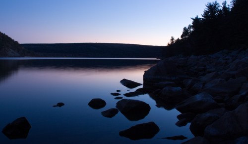 A tranquil lake at dusk, with smooth water reflecting the twilight sky and rocky shores lined with trees.