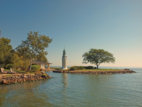 A serene lakeside scene featuring a lighthouse, a tree, and a small building under a clear blue sky.