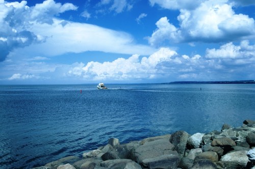 A serene seascape with a boat on calm waters, surrounded by blue skies and fluffy clouds, with rocky shore in the foreground.