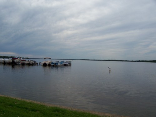 A calm lake scene with boats docked at a marina under a cloudy sky.