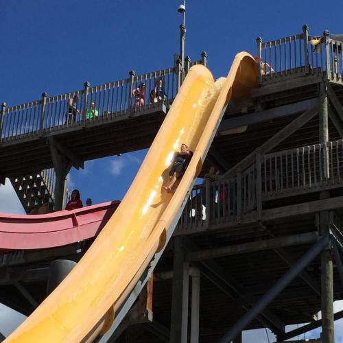 A person slides down a large yellow water slide at a water park, with spectators on the platform above.