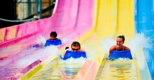 Three people on blue mats slide down colorful water slides, splashing into the water below.