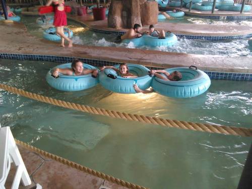 Three children float on blue inner tubes in a lazy river, while others play in the water nearby.