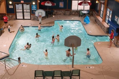 Indoor pool scene with children and adults swimming, playing, and relaxing around the water. Poolside chairs visible.