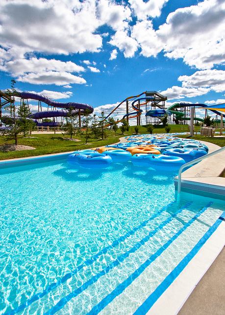 A clear blue pool with colorful inner tubes, surrounded by green grass and water slides under a bright, cloudy sky.