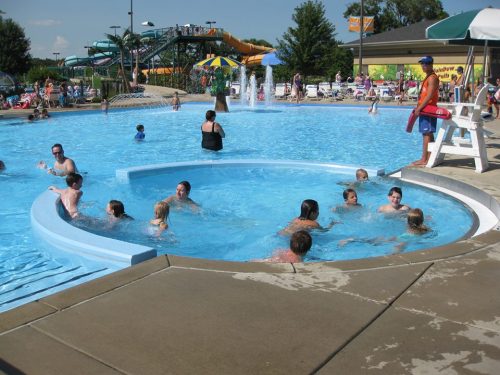 A busy water park pool with children swimming, a lifeguard on duty, and water slides in the background.